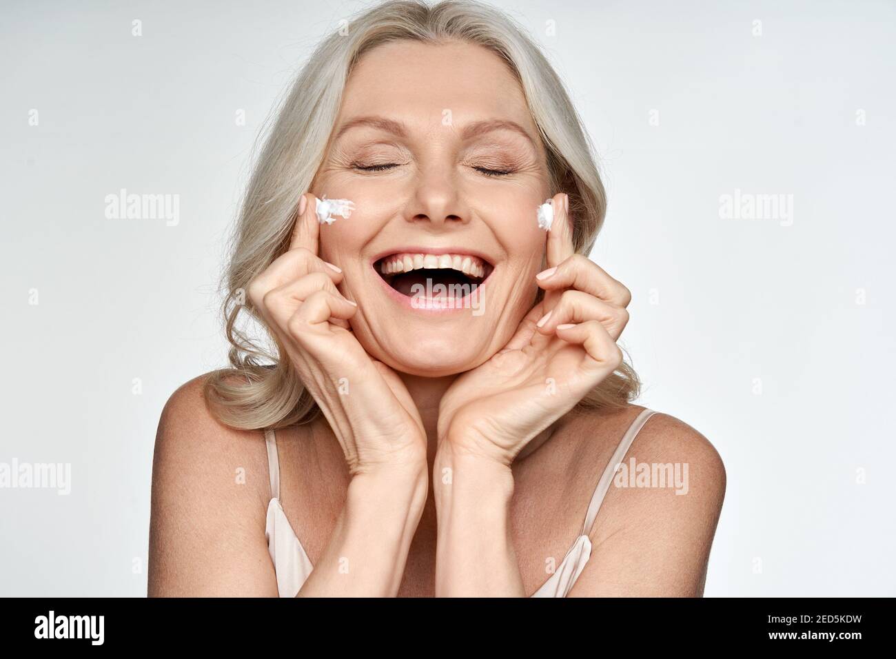Happy funny 50s middle aged woman applying cream on face isolated on white. Stock Photo