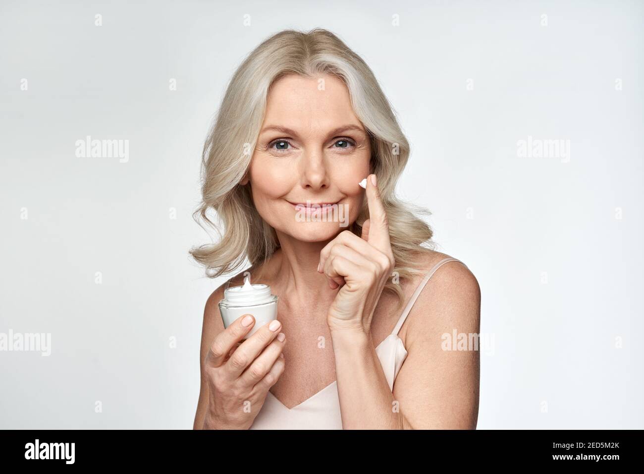Smiling 50s middle aged woman putting facial cream on face looking at camera. Stock Photo