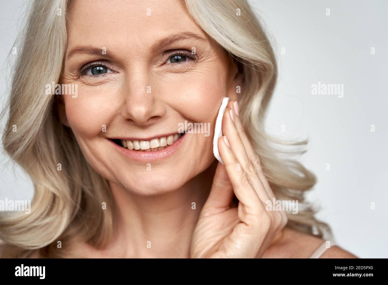 Smiling 50s middle aged mature woman holding cotton pad cleaning face skin. Stock Photo