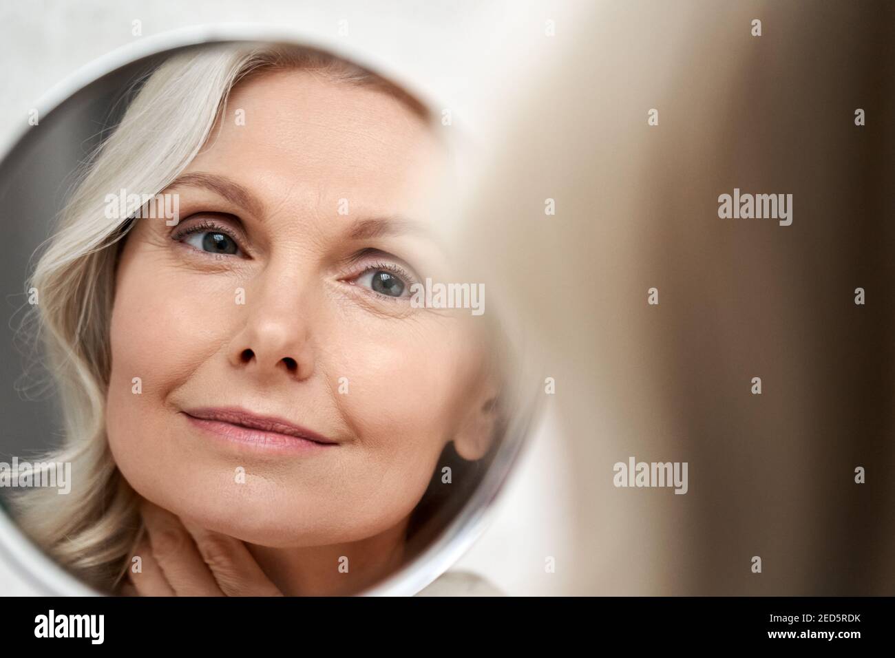 Happy 50s middle aged woman touching face skin looking in mirror reflection. Stock Photo