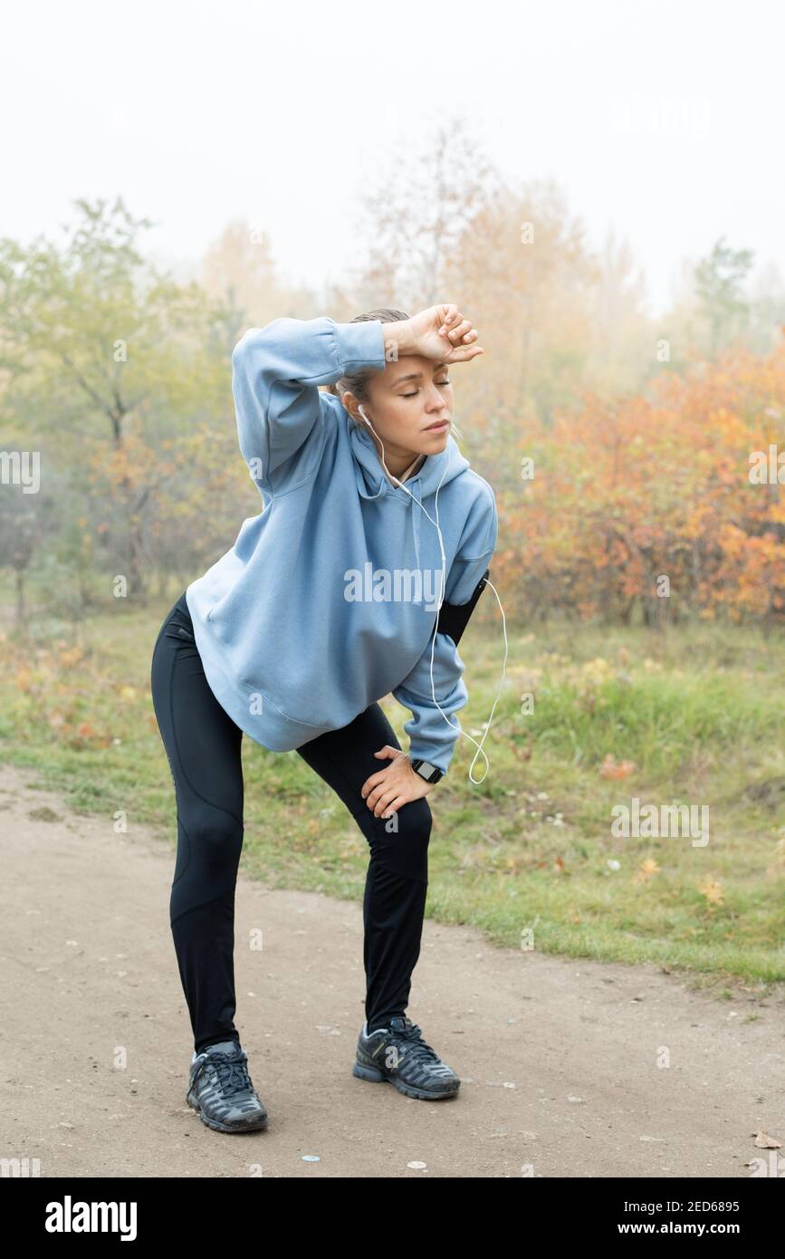 Young tired sportswoman in activewear bending slightly forwards while standing with bent knees on path after training and touching forehead Stock Photo