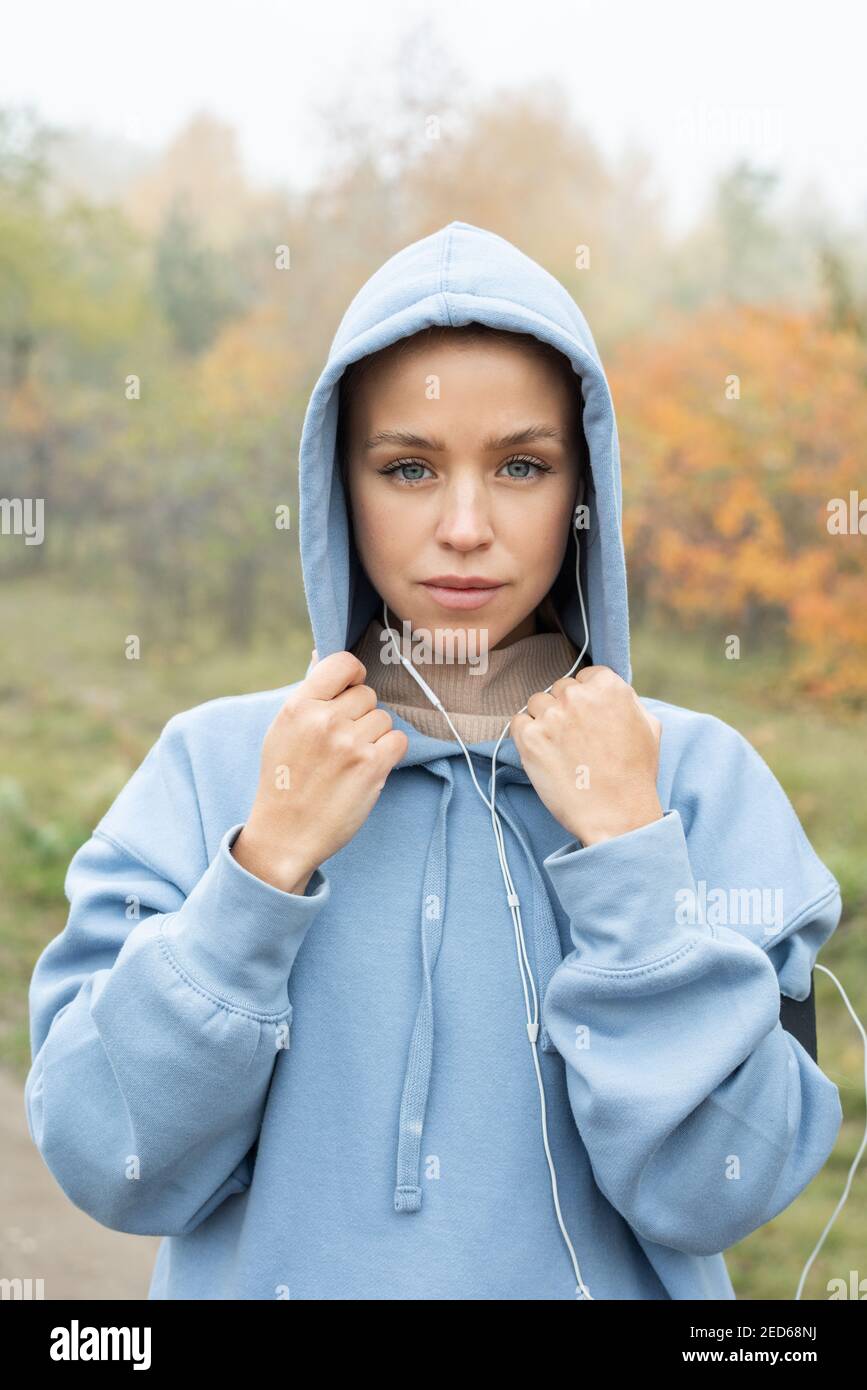 Young sportswoman in earphones and blue hoodie standing in front of camera against trees in park and looking at you after outdoor training Stock Photo