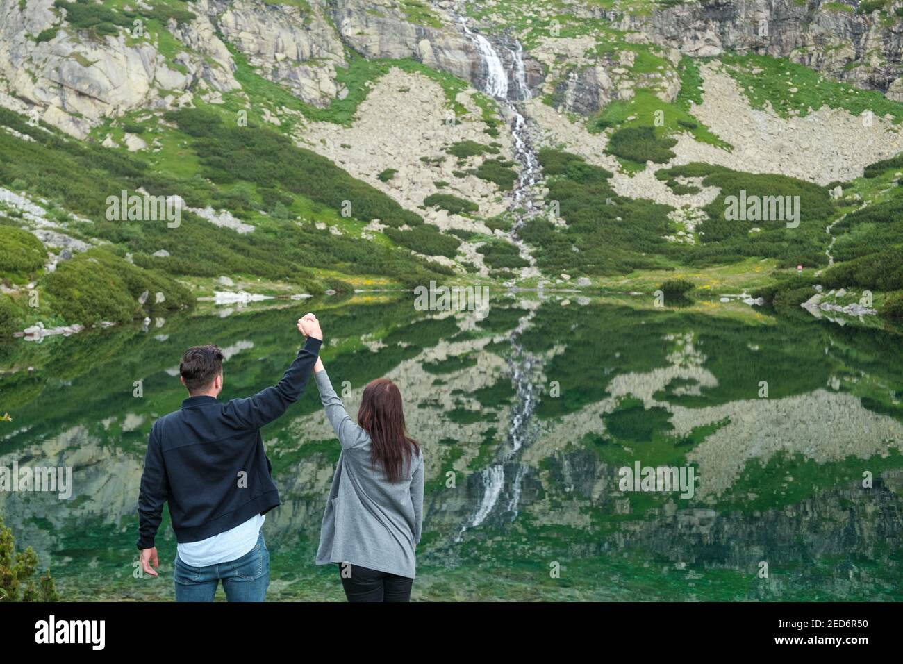 Heterosexual couple holding hands against amazing view of waterfall and lake with transparent clear water. Stock Photo