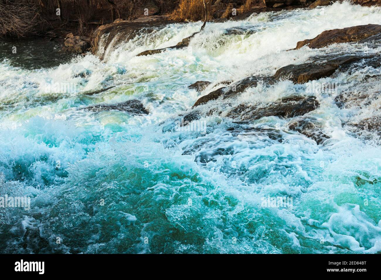 A seething waterfall spills through the rocky valley at the winter. Water splashes with foam on the waves. Stock Photo