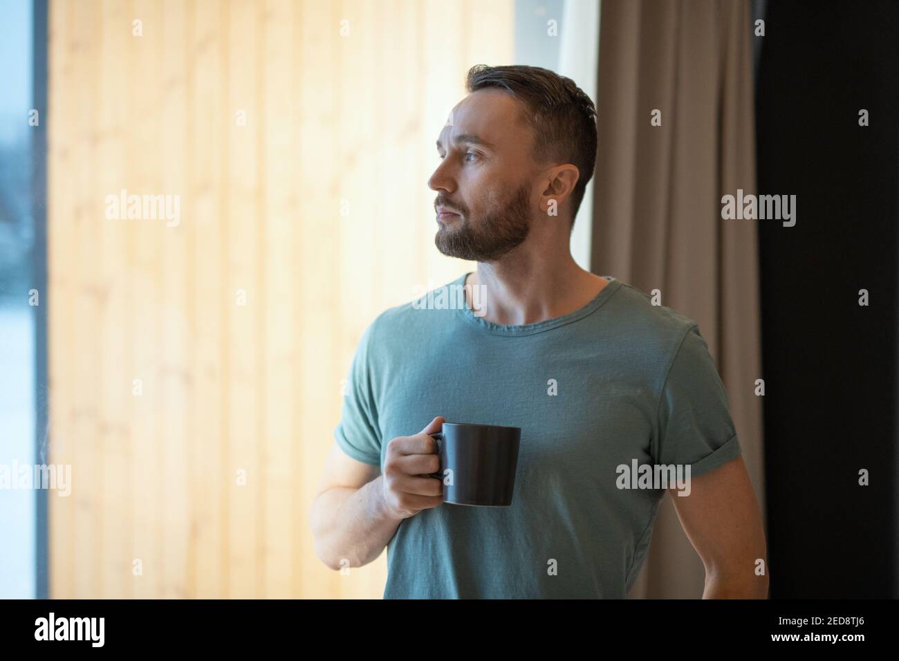 Young bearded relaxed man in grey t-shirt holding mug with hot tea or coffee and looking through large window while staying at home Stock Photo