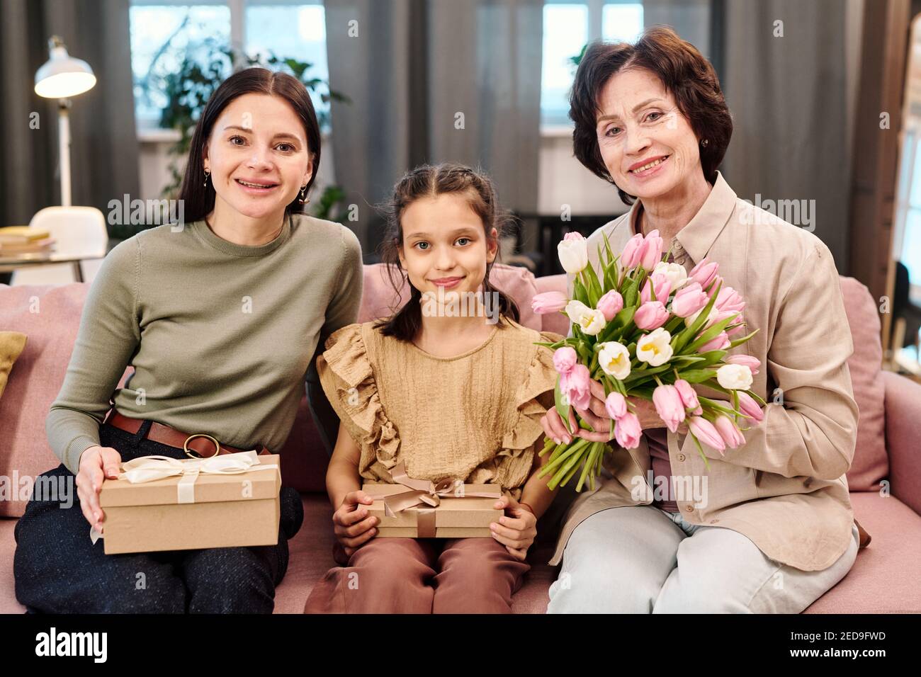 Two cheerful females in casualwear and adorable little girl holding packed giftboxes with presents and bunch of gresh tulips while sitting on couch Stock Photo