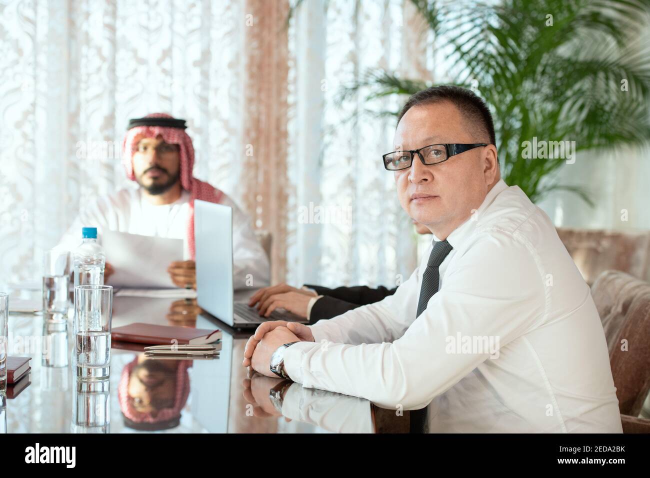 Serious mature business delegate in eyeglasses and formalwear looking at you while sitting by table against his secretary and Arab partner Stock Photo