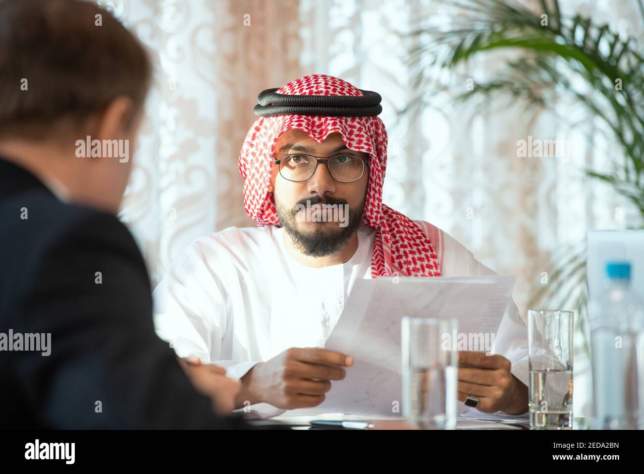 Young serious Arab delegate in eyeglasses and national clothes reading contract of business partnership and looking at mature businessman Stock Photo