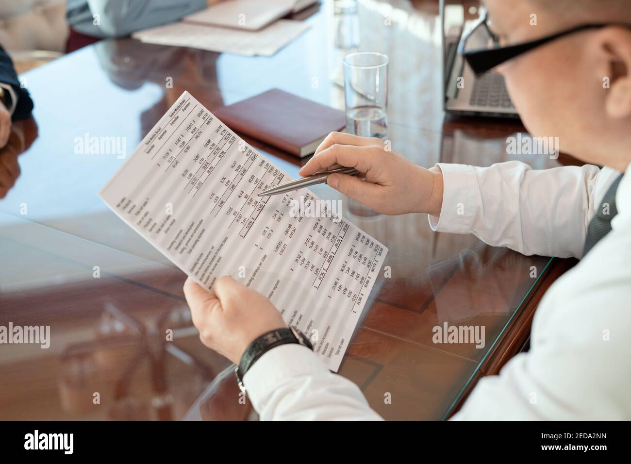 Mature delegate in formalwear and eyeglasses bending over table while looking through document with financial data during negotiation Stock Photo