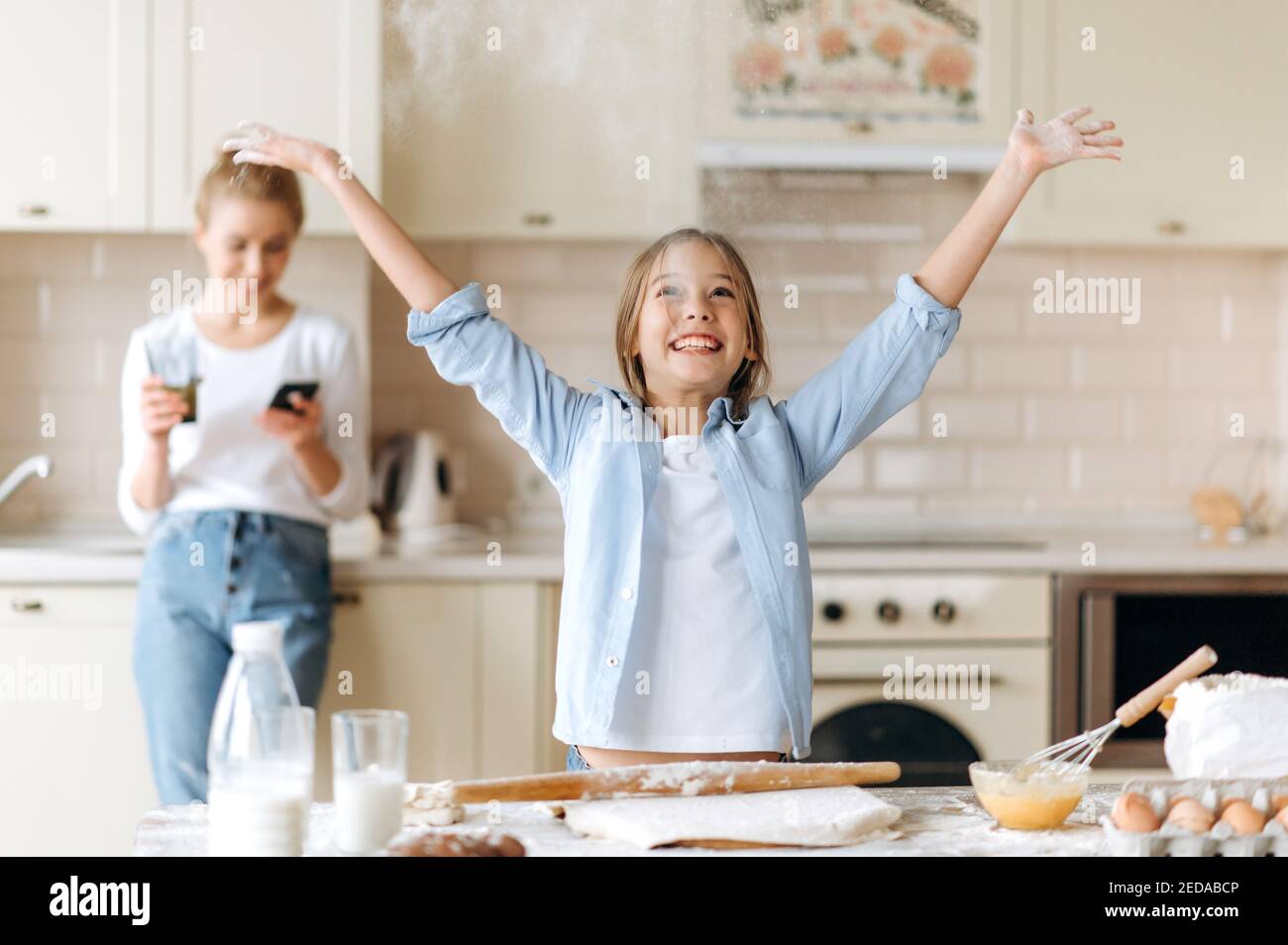 Little cook. A cute caucasian little girl learns to cook a pie, indulges in flour, smiling, mom stands on the background using the phone Stock Photo