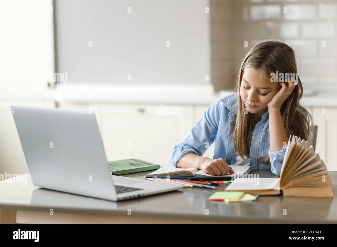 Schoolgirl is tired from online lesson. Distance learning during quarantine. Focused caucasian pretty schoolgirl doing her homework at home using laptop, sitting at the work desk Stock Photo