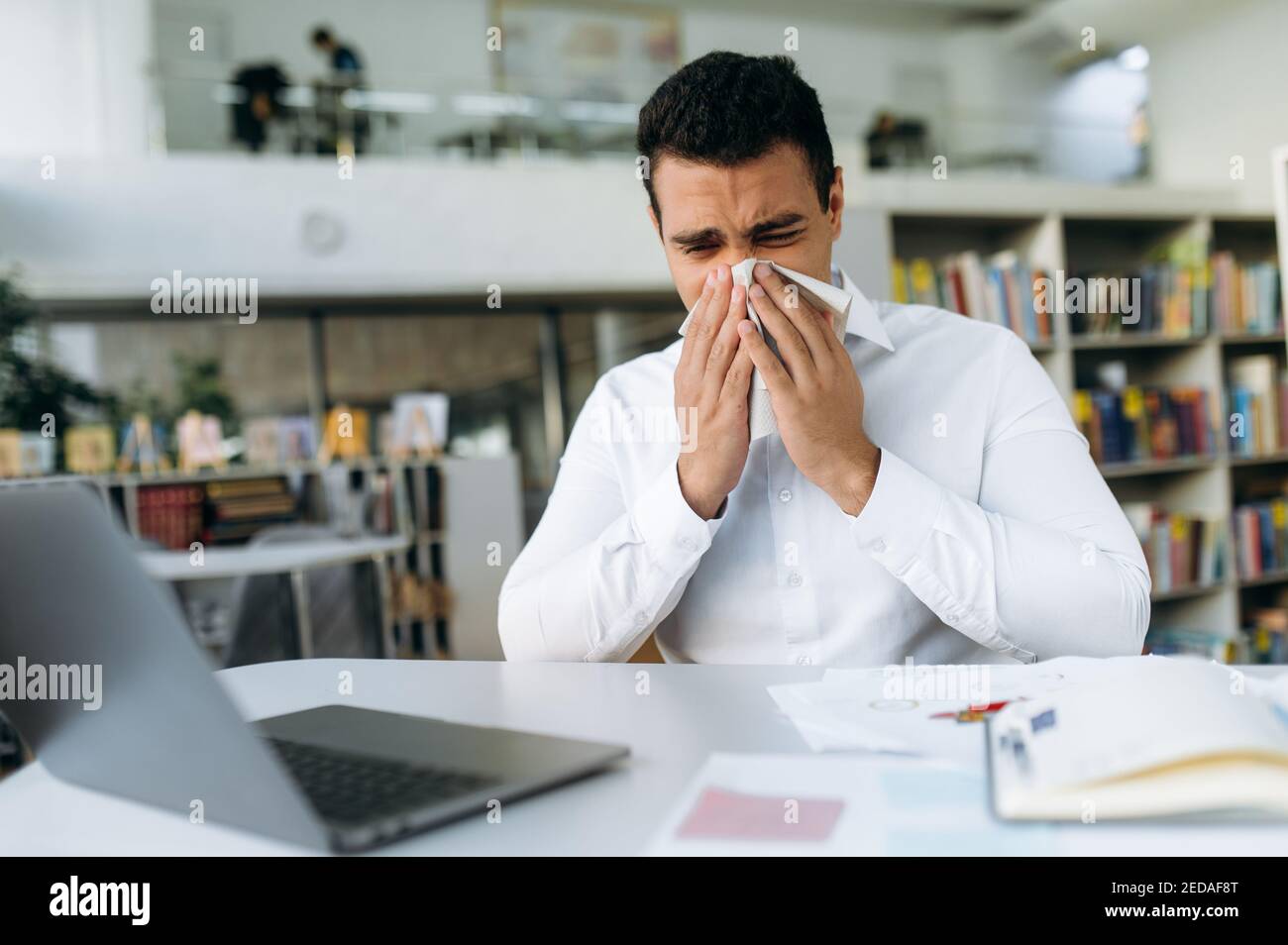 Unhealthy male employee sit at the desk, using a paper tissue, sneezing. Business man feeling unwell, struggling with running nose. Hispanic guy remotely working or studying, while sickness Stock Photo