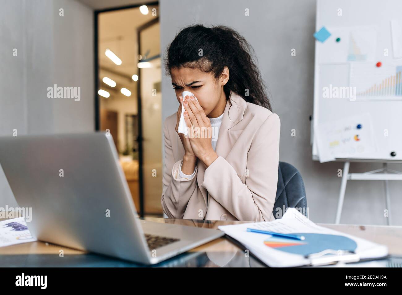Business woman sit at the desk, using paper tissue, sneezing. Beautiful female employee feeling unwell, sick with running nose. African american lady remotely working or studying, while sickness Stock Photo