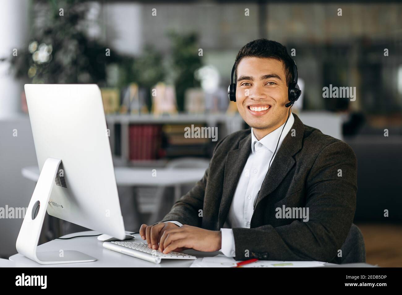 Support service, help desk. Portrait of a young successful hispanic call center worker, manager or entrepreneur sitting at work desk wearing headphones looking at the camera with a smile Stock Photo