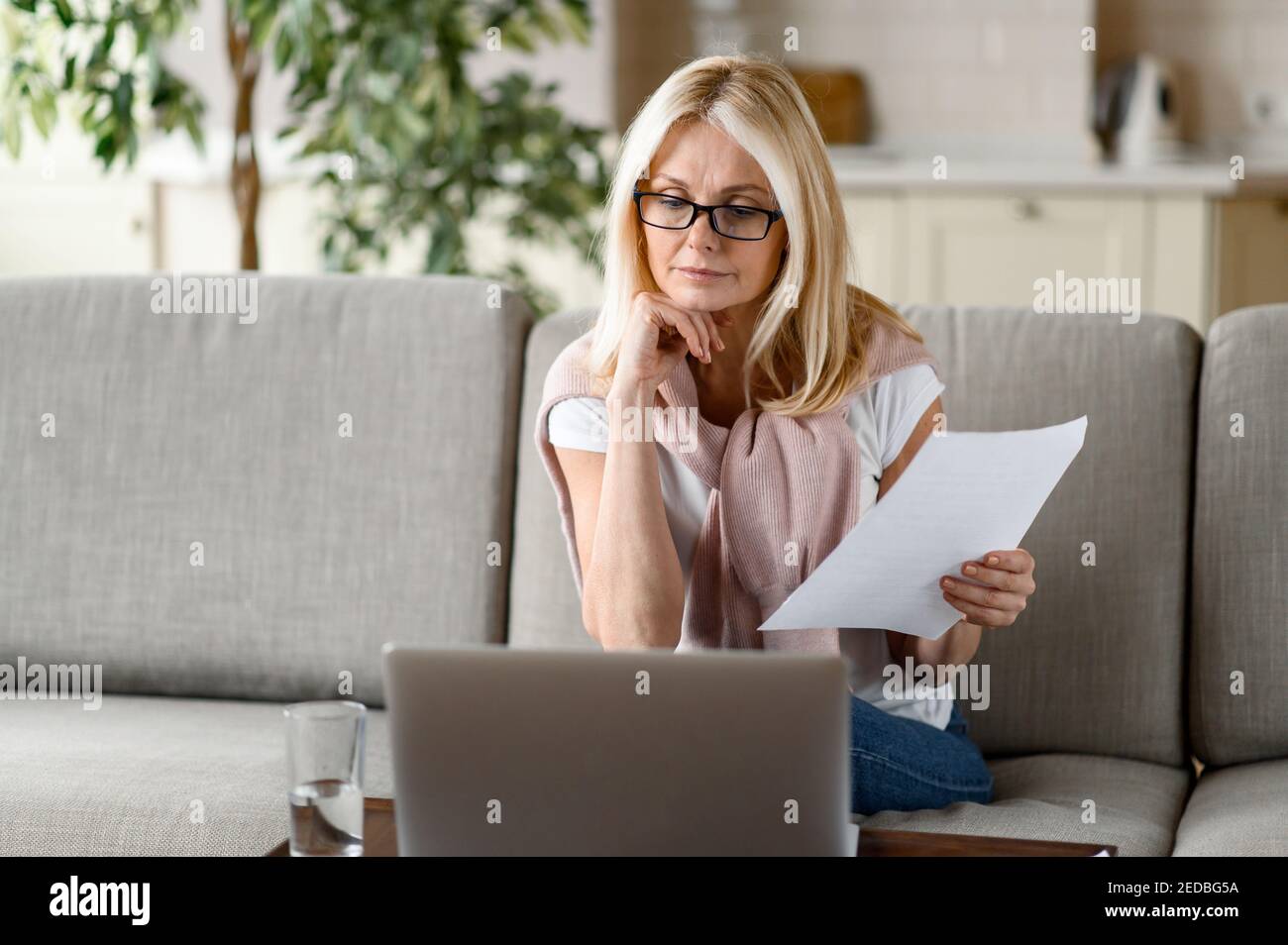 Focused senior blonde works at home remotely, sitting on the couch in the living room, analyzes monthly reports, uses a laptop. Work from home Stock Photo