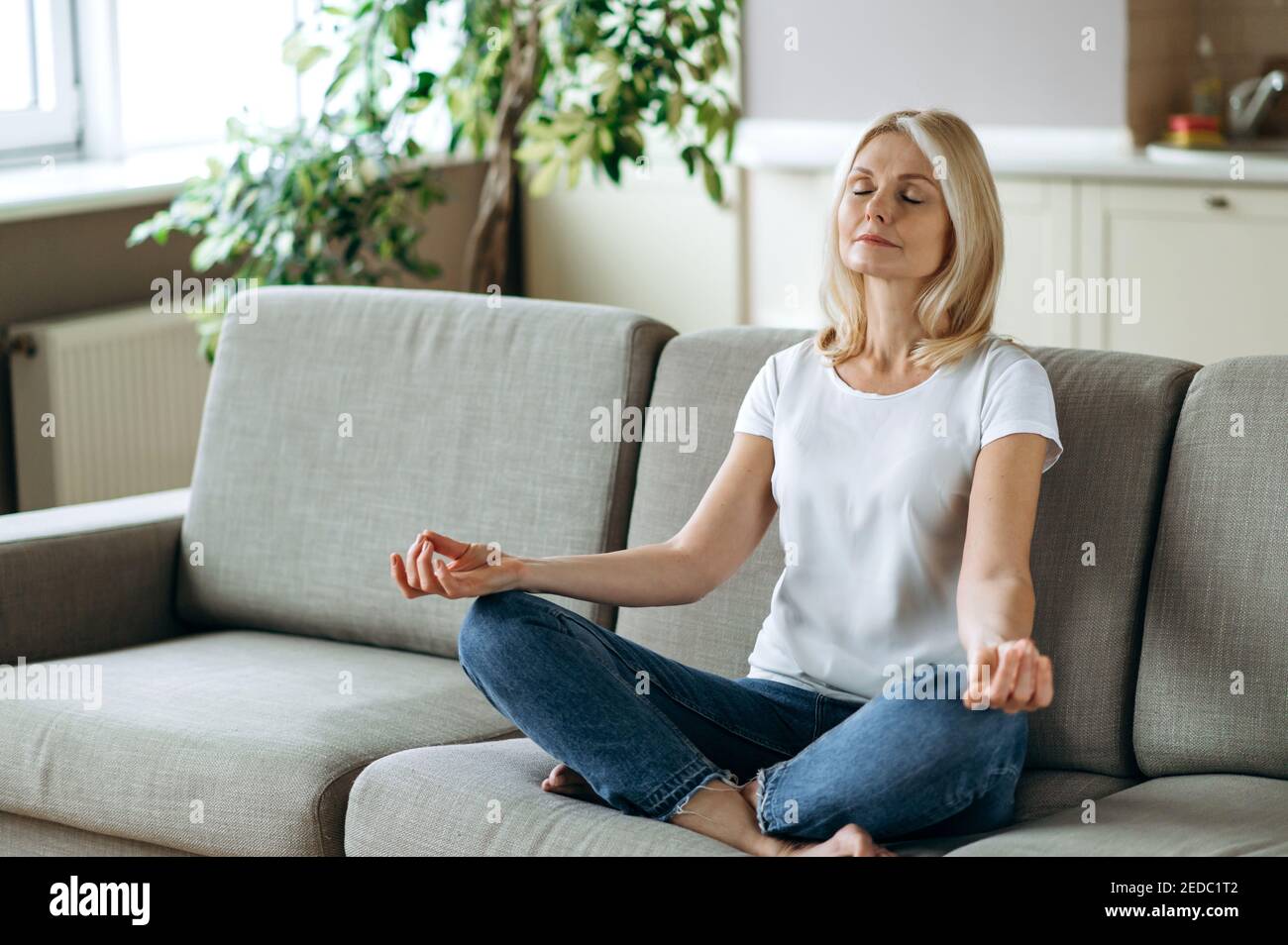 Satisfied attractive middle aged female sits on sofa at home and chilling. Blonde mature is practise yoga and meditation in lotus position with eyes closed, relieve stress, calmness concept Stock Photo