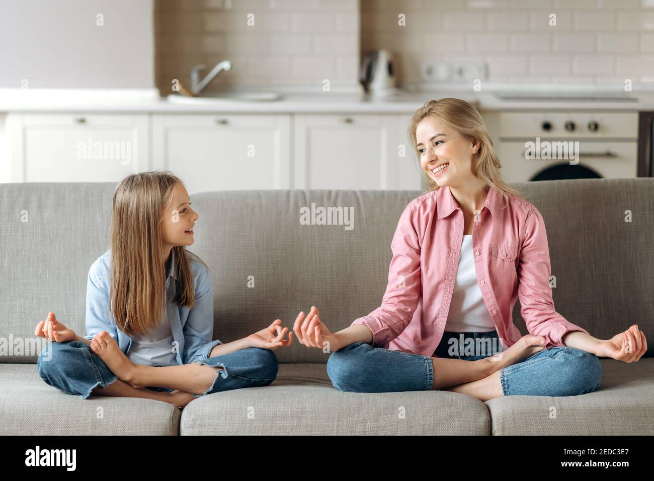 Happy young caucasian mom with beloved little daughter doing yoga and meditation at home on the couch, sitting in lotus position, calmness, relaxing, looks at each other Stock Photo