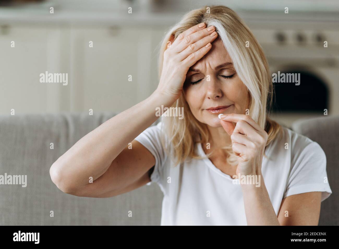 Upset unhealthy caucasian mature woman sitting at home on sofa drinking pain relieving pill for headache with eyes closed Stock Photo