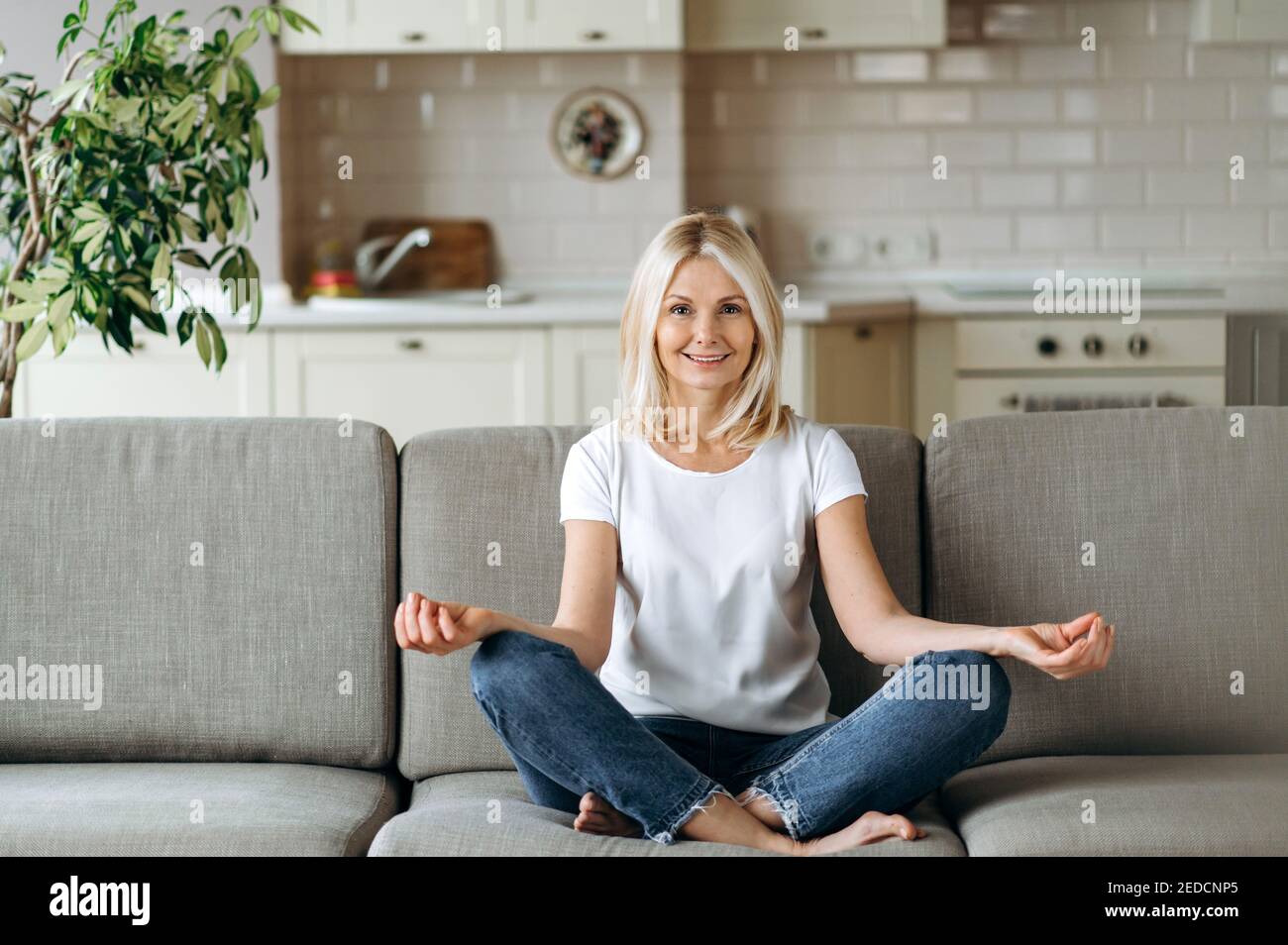 Smiling caucasian middle aged female sits on sofa at living room and practise yoga, meditation in lotus position looks at camera and smiling, relieve stress Stock Photo
