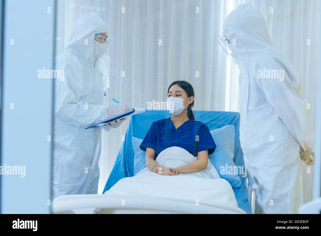 Covid-19 doctors in ppe suit with face shield and mask checking and treating young woman patient lying on bed suffering from coronavirus undergoing routine checkup Stock Photo
