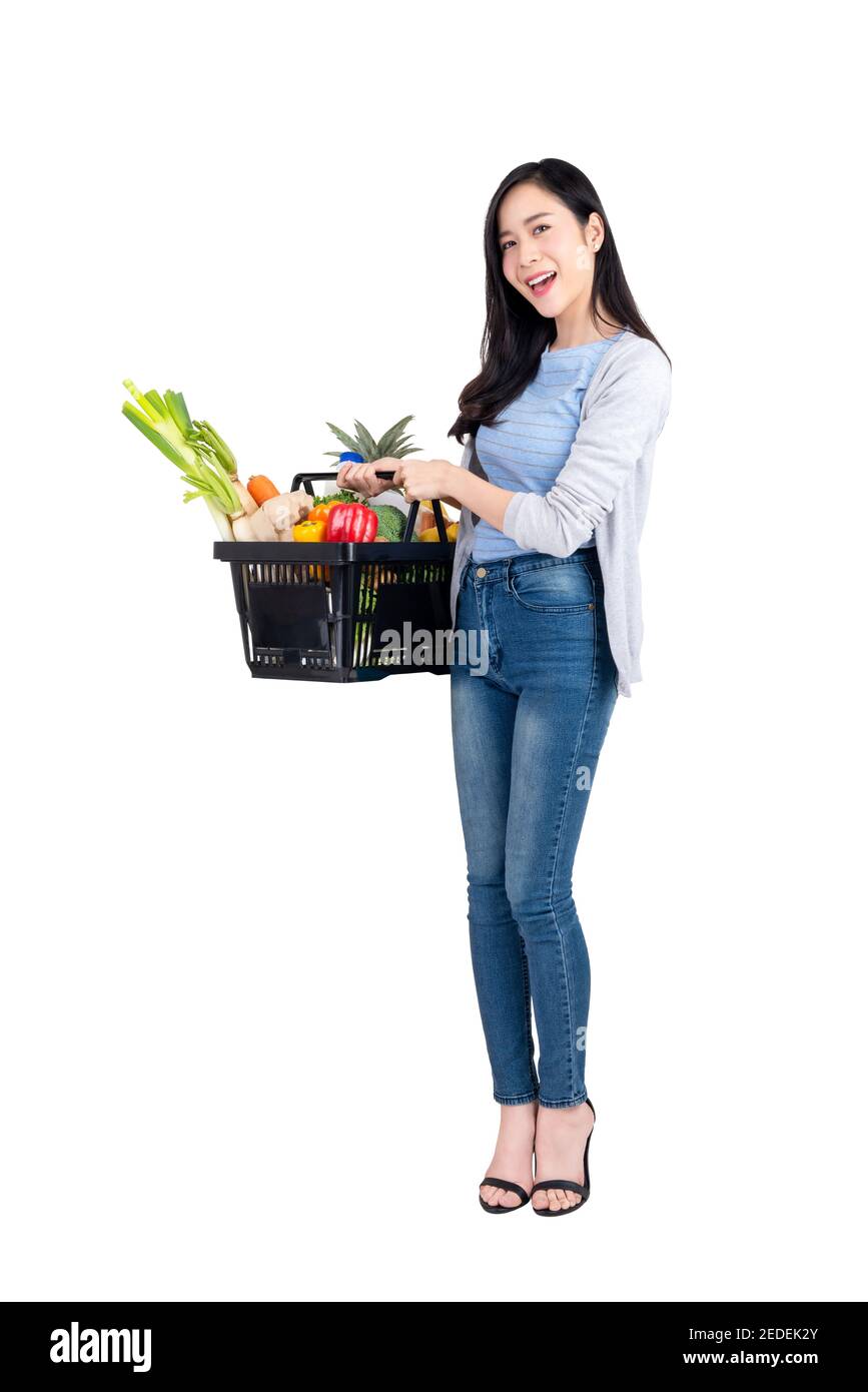 Beautiful Asian woman holding supermarket shopping basket full of vegetables and groceries, studio shot isolated on white background Stock Photo