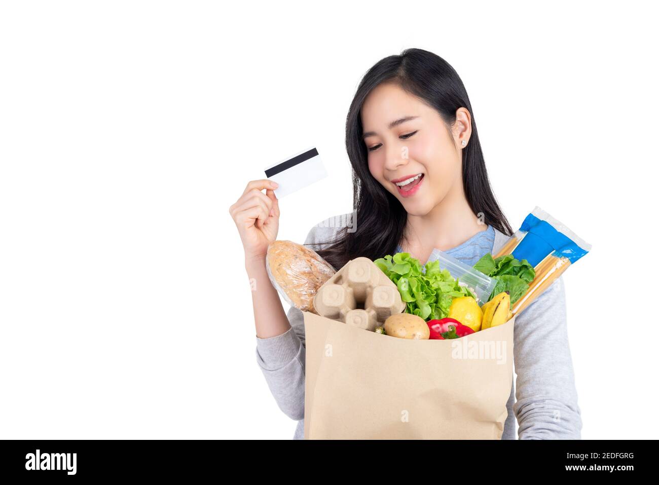 Beautiful Asian woman holding paper shopping bag full of vegetables and groceries just bought with credit card, studio shot isolated on white backgrou Stock Photo
