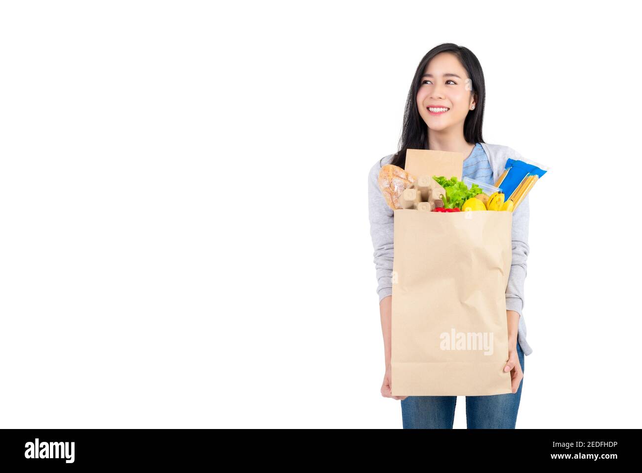 Beautiful Asian woman holding paper shopping bag full of vegetables and groceries looking at the space aside, studio shot isolated on white background Stock Photo