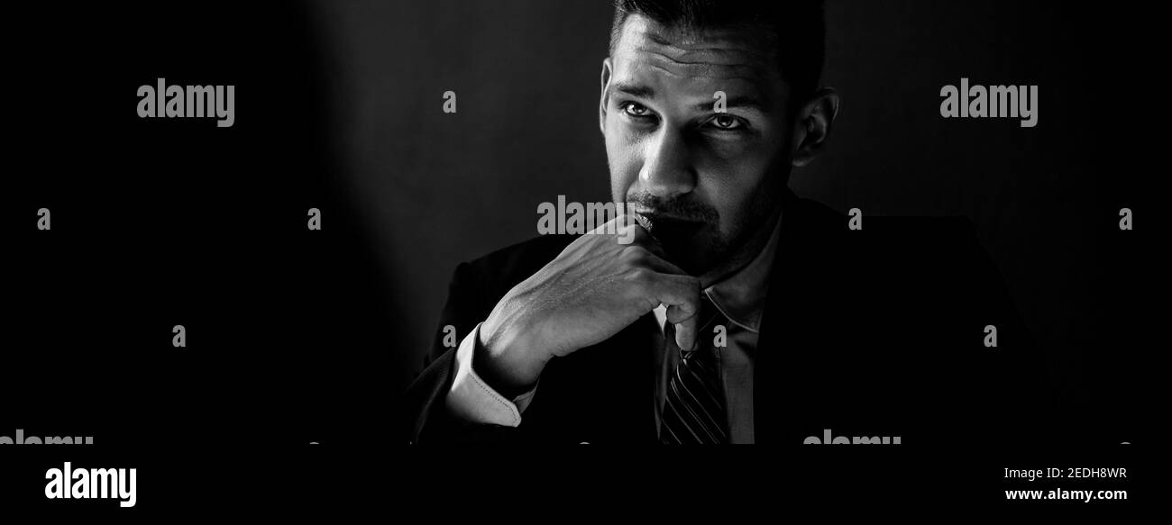 Black and white tone banner of interrogator in shadow with serious face thinking and staring at criminal man in interrogation room Stock Photo