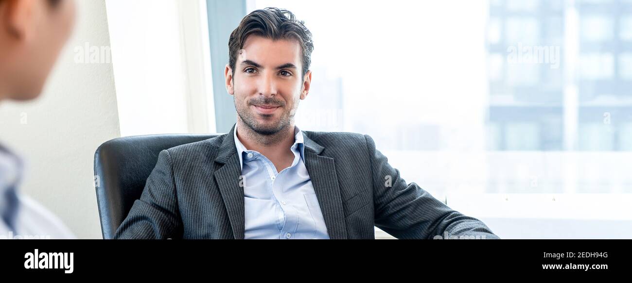 Handsome Hispanic businessman as a boss sitting in a chair listening to his colleague in office banner background Stock Photo