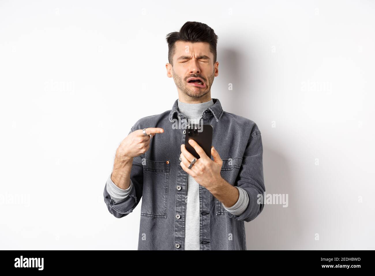 Sad crying guy pointing at smartphone and sobbing, complaining or feeling miserable, standing on white background Stock Photo