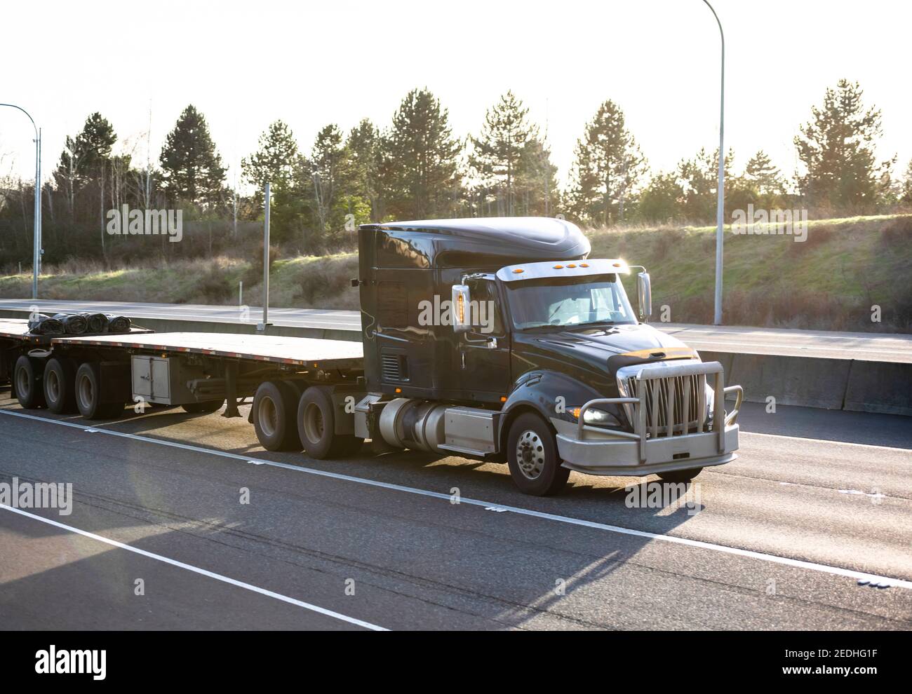 Big rig industrial classic black powerful semi truck tractor with low cabin transporting two empty flat bed semi trailers running on the highway road Stock Photo