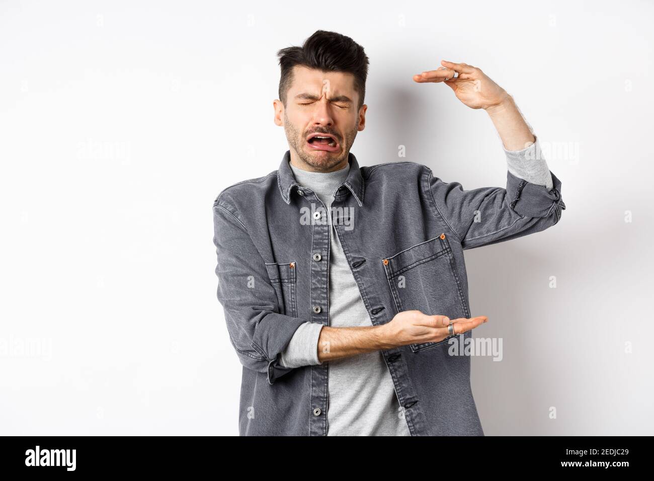 Sad man crying and showing big size object, shaping large thing and complaining, sobbing while standing on white background Stock Photo