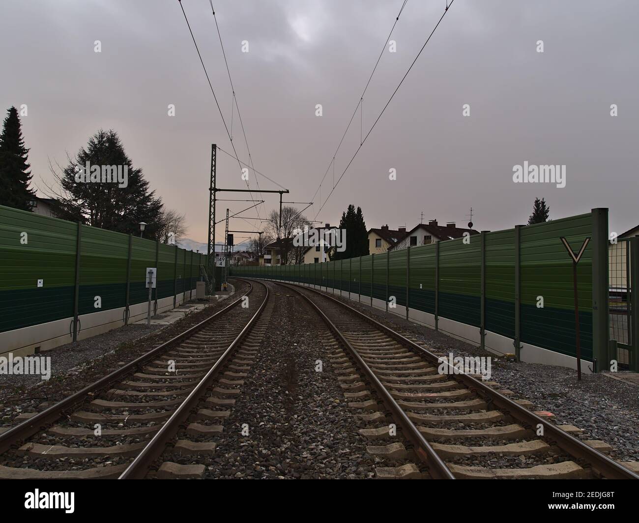 Two-lane railroad tracks with overhead cable and green colored noise protection barrier in a village on cloudy winter day with diminishing perspective. Stock Photo