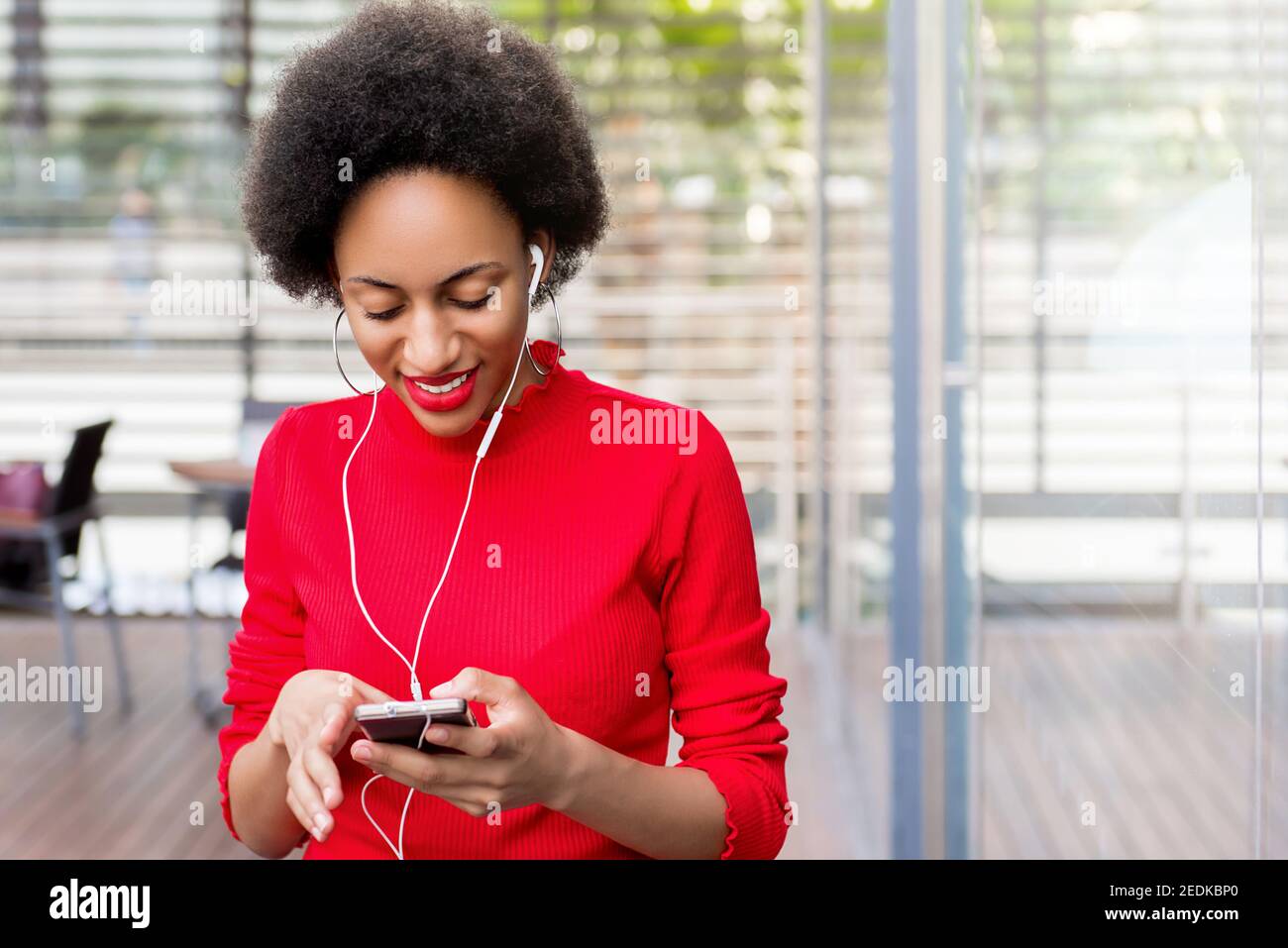 Young beautiful afro black woman wearing earphones listening to musics online via smartphone device while sitting in cafe Stock Photo