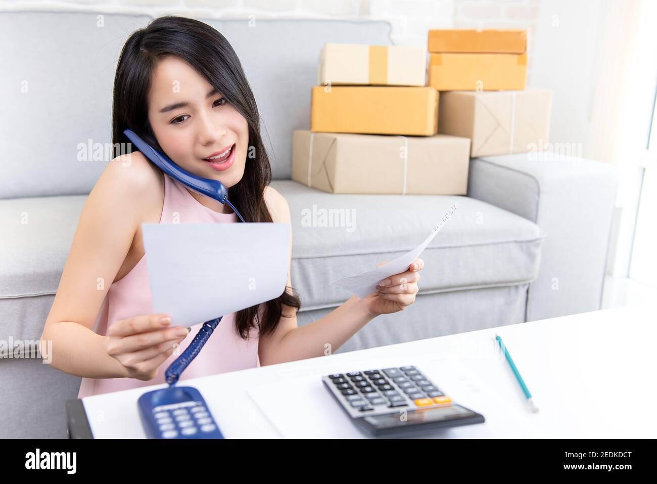 Young Asian woman entrepreneur or freelance online seller working at home confirming orders from customer on the phone preparing for delivery Stock Photo