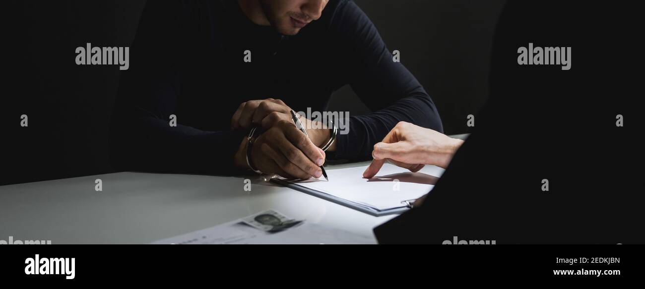 Criminal man with handcuffs signing document in interrogation room after committed a crime Stock Photo