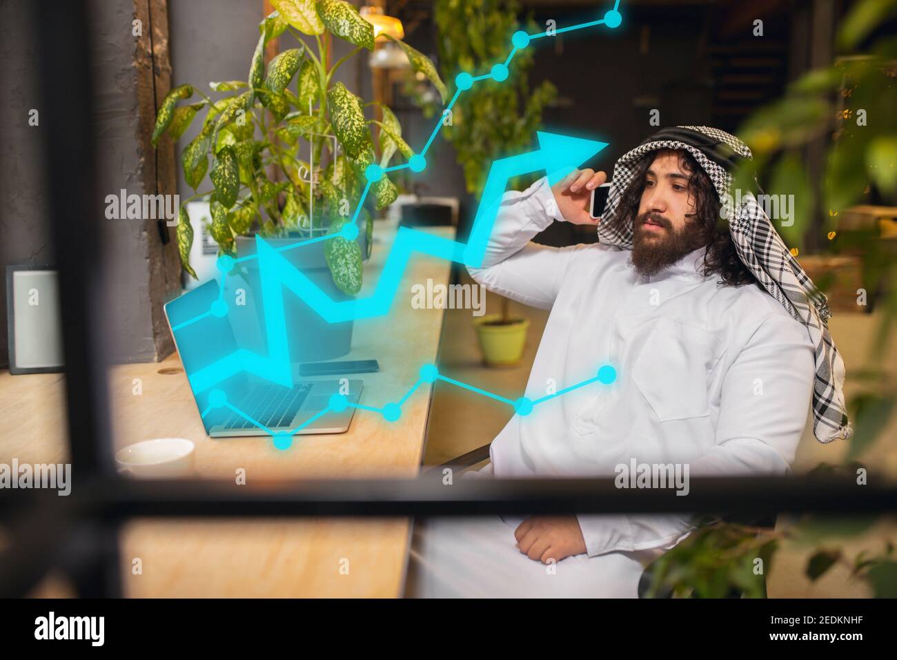 On the phone. Arabian businessman working in office, business centre using device, gadget. Modern saudi lifestyle. Man in traditional wear looks confident, busy. Ethnicity, finance, income uncrease Stock Photo