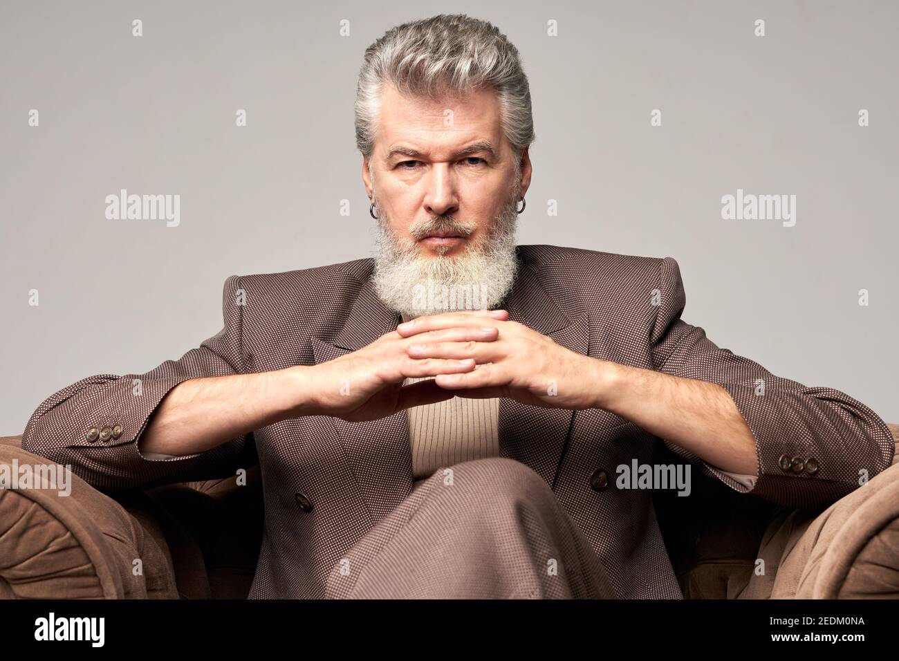 Portrait of brutal mature businessman with beard wearing elegant suit looking at camera, sitting with hands crossed in armchair while posing isolated over light background. Front view Stock Photo