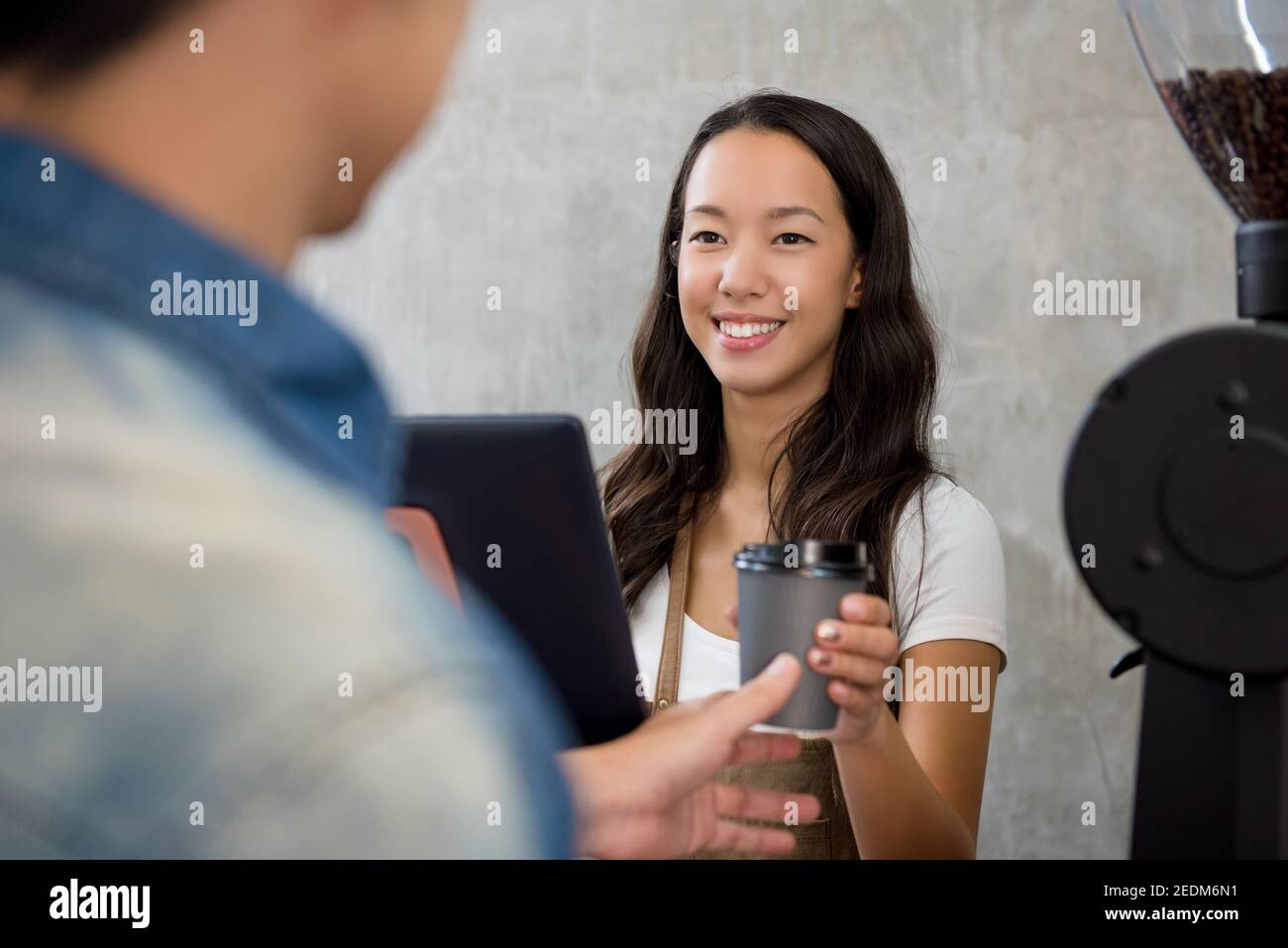 Young friendly Asian woman staff giving a cup of takeaway coffee to customer at counter in cafe Stock Photo
