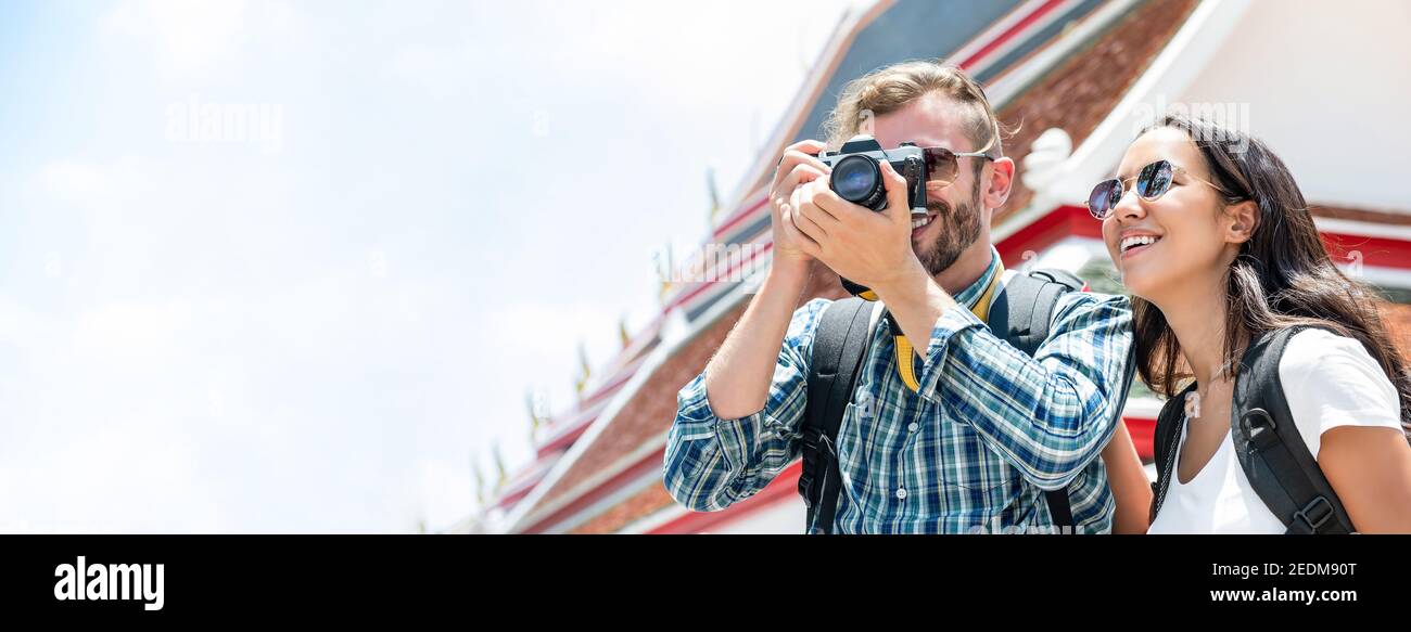 Interracial tourist couple taking photos during summer holiday trip in Bangkok Thailand with Thai temple in background, panoramic banner Stock Photo
