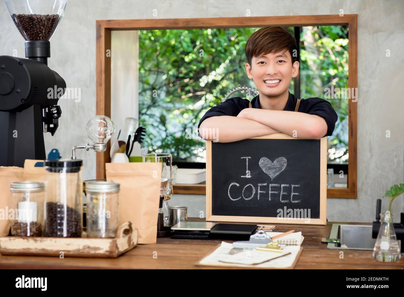 Cheerful young Asian man entrepreneur at counter in his own cafe with I love coffee sign Stock Photo