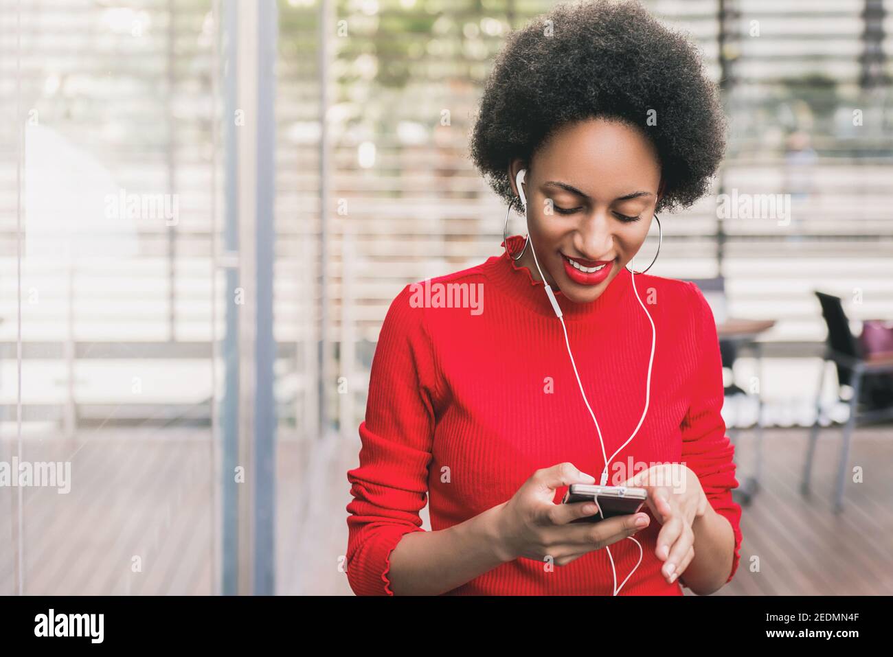 Young beautiful afro black woman wearing earphones listening to musics online via smartphone device while sitting in cafe Stock Photo