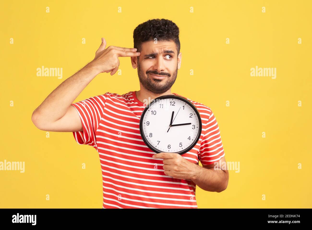 Kill me please. Busy unhappy bearded man in striped t-shirt holding fingers near temple like gun going to make shot holding big wall clock, deadline. Stock Photo