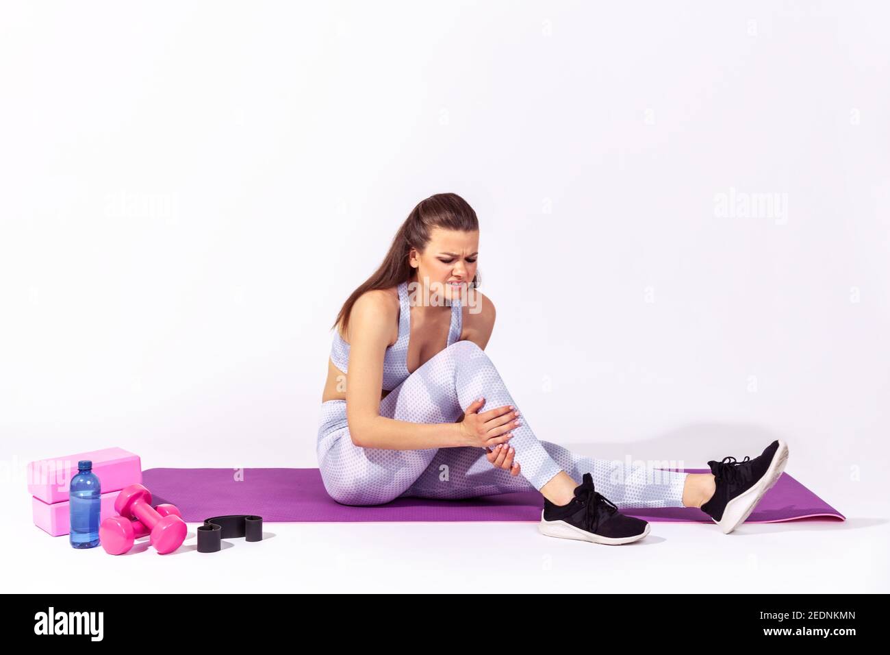 Full length athletic woman in white sportswear sitting on gym mat touching injured caviar, suffering calf muscle strain after yoga workout. Indoor stu Stock Photo