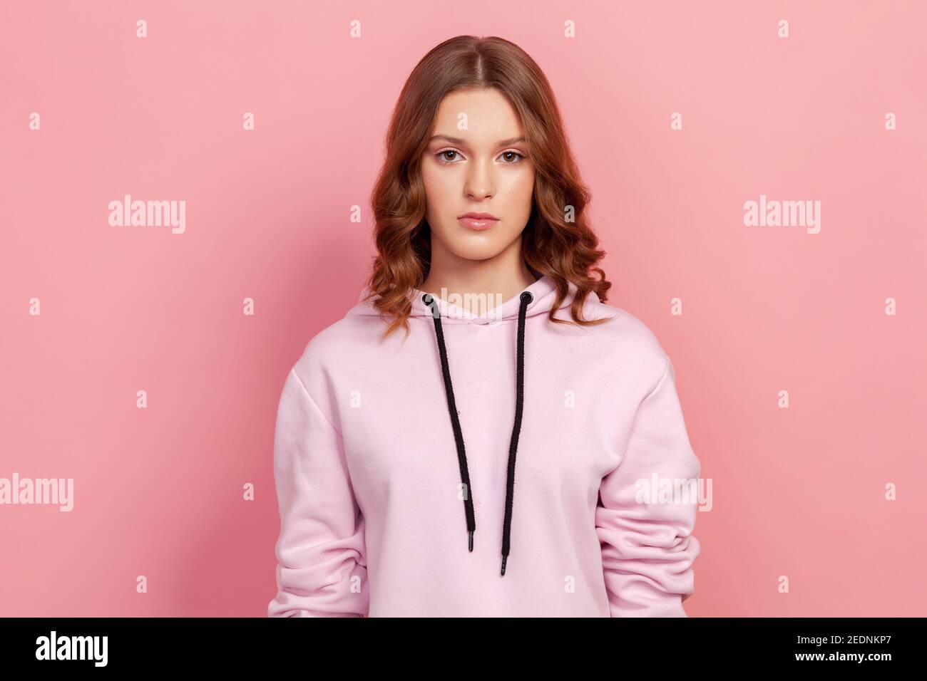 Portrait of serious brunette teenager girl in hoodie attentively looking at camera. Indoor studio shot, isolated on pink background Stock Photo