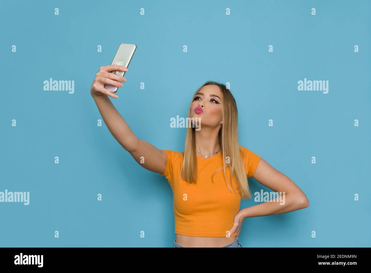 Cute woman in orange top is holding telephone, taking a selfie and sending a kiss. Waist up studio shot on blue background. Stock Photo