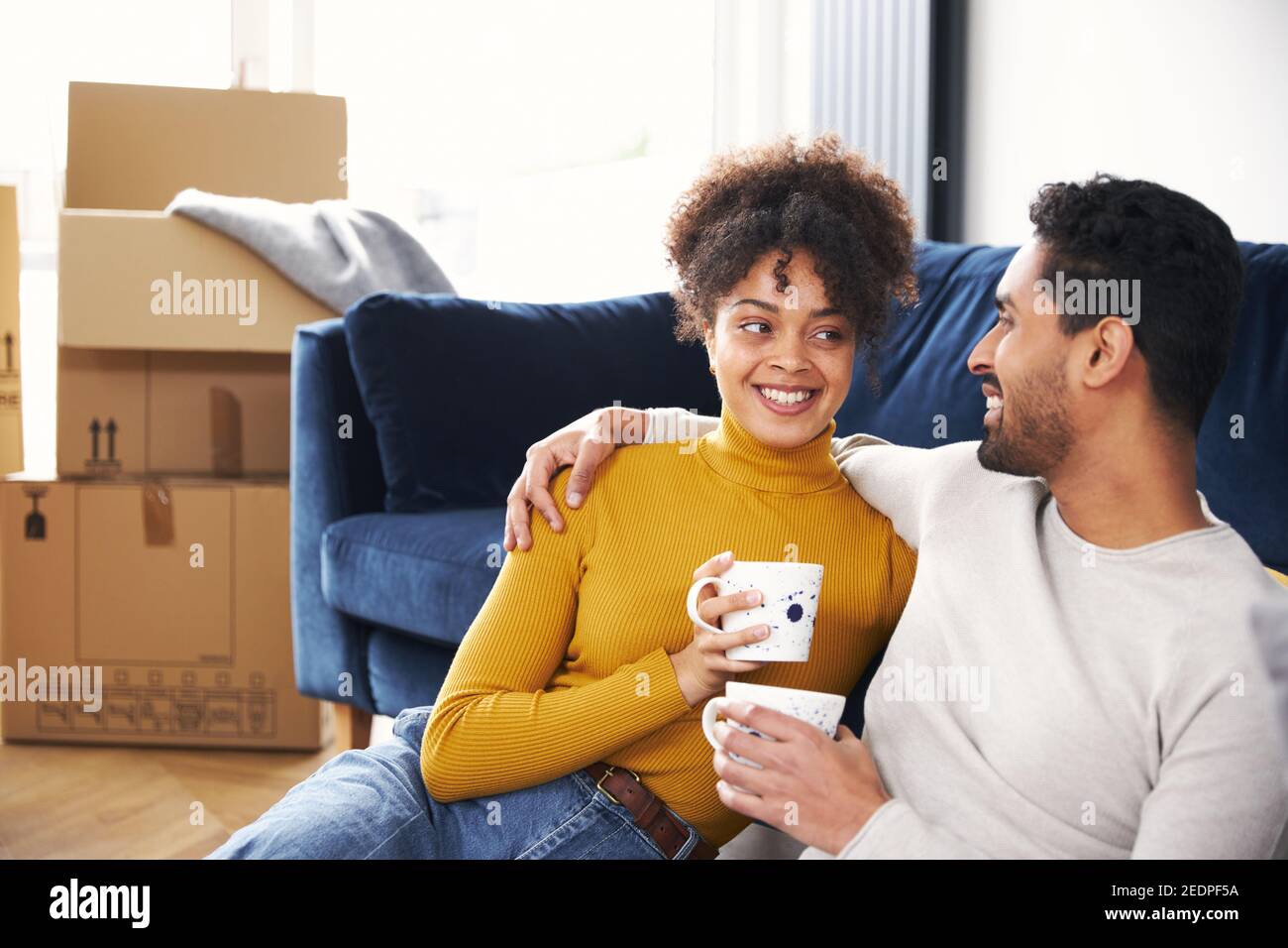 Young mixed ethnicity couple taking a break on moving day into new home sitting on floor in lounge drinking coffee surrounded by removal boxes Stock Photo