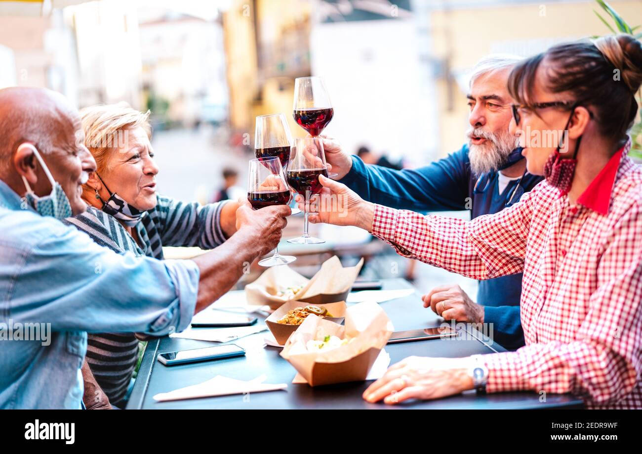Senior friends toasting red wine at winery bar dehor with open face mask - New normal lifestyle concept with happy retired couples having fun together Stock Photo