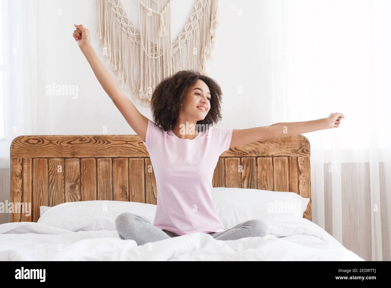 Happy teen african american fresh vitality lady sits on bed, stretching arms after sleep and enjoying morning Stock Photo