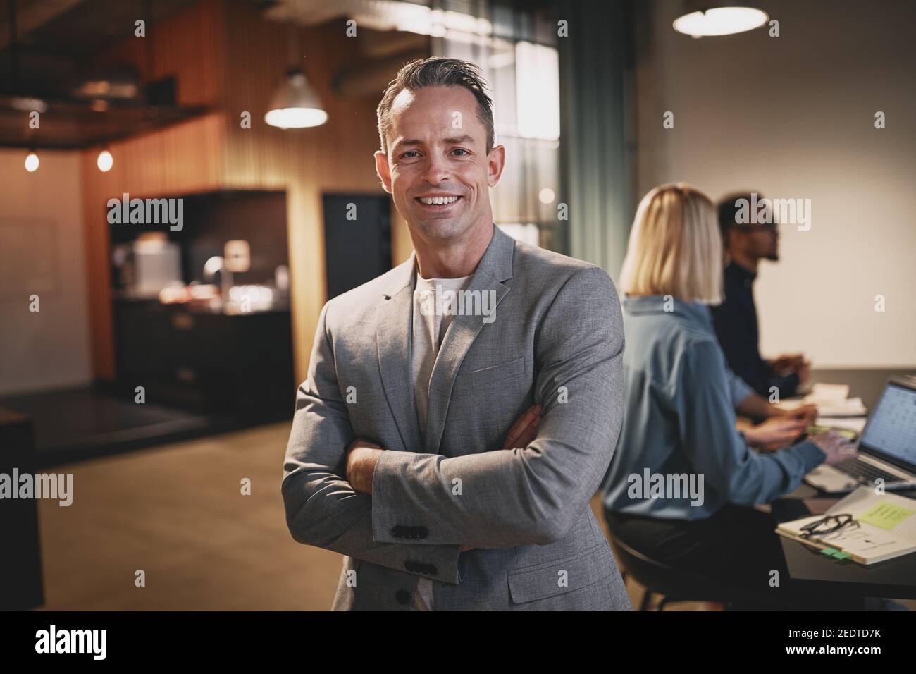 Smiling mature businessman standing with his arms crossed in an office with colleagues talking at a table in the background Stock Photo